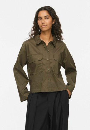 Femme aux longs cheveux bouclés porte une chemise boutonnée vert foncé avec deux poches poitrine et un pantalon noir ample, debout avec les mains dans les poches.