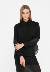 Camicia nera con bottoni, maniche trasparenti, ricami floreali sulle spalle e colletto classico. Texture liscia con dettagli sottili.