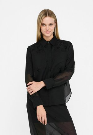 Camicia nera con bottoni, maniche trasparenti, ricami floreali sulle spalle e colletto classico. Texture liscia con dettagli sottili.