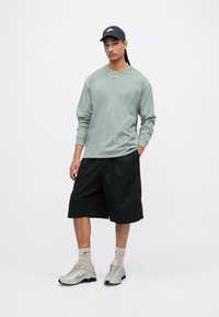Maglietta verde a maniche lunghe con scollatura rotonda, pantaloni corti neri, scarpe da ginnastica grigie e cappellino nero; presenta il logo Nike sulla maglietta e sulle scarpe.