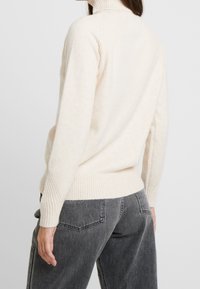 Kobieta w kremowym swetrze z golfem i ciemnoszarych, wysokich dżinsach, pokazana od tyłu, na jednolitym tle.