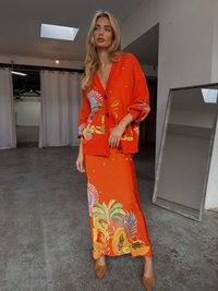 Cardigã laranja com padrões coloridos de flores e frutas, combinado com uma saia longa de cetim laranja com designs tropicais. Acessorizado com sapatos de salto bege.