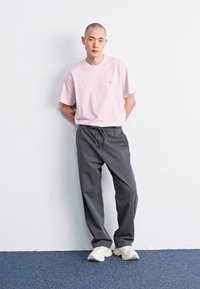 T-shirt rose clair avec un petit logo, associée à un pantalon de survêtement gris foncé et des baskets blanches, se tenant devant un mur uni.
