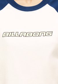 Λευκό και ναυτικό μπλε raglan μπλουζάκι με το λογότυπο "BILLABONG" σε έντονη, περιγραμμένη γραμματοσειρά στο κέντρο του στήθους.