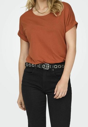 Femme portant un t-shirt manches courtes couleur rouille rentré dans un jean noir taille haute avec une ceinture noire à œillets métalliques.