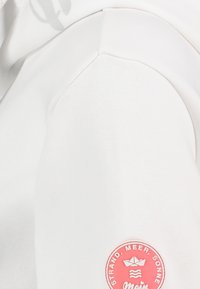 Weißer Sweatshirt mit weicher Textur, einem runden pinken Logo-Patch mit nautischem Design am linken Ärmel. Einfaches, lässiges Design.