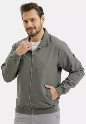 Brodaty mężczyzna poprawiający kołnierz szarej kurtki na zamek, z jedną ręką w kieszeni, stojący na jednolitym tle.