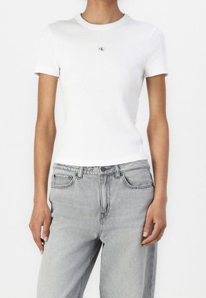 Žena nosí bílý přiléhavý žebrovaný top s krátkými rukávy a malým logem Calvin Klein na hrudi a světle šedé džíny s vysokým pasem.