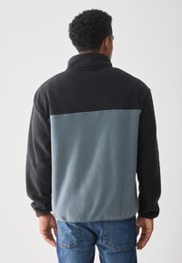 Chaqueta de forro polar con cuello alto; presenta un diseño a bloques de color en negro y gris azulado. Textura suave; mangas largas; ajuste relajado.