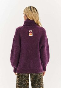 Fialový, veľký pletený sveter s vysokým golierom a štruktúrovanou látkou. Na chrbte má malú etiketu. Kombinovaný s nohavicami s potlačou.