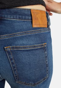 Los pantalones vaqueros de denim azul presentan un acabado texturizado, costuras naranjas y una etiqueta de cuero color marrón claro en la cintura con detalles de texto en relieve.
