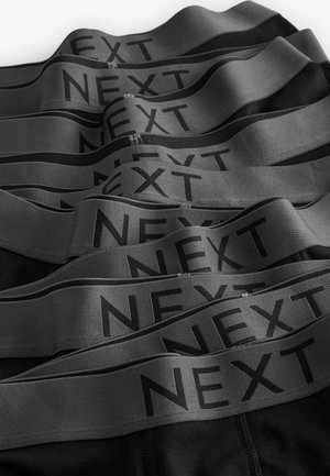 Crne gaćice s širokim sivo-elastičnim pojasom na kojem je u crnom tisku napisano "NEXT". Više pari složenih jedan na drugi, prikazujući teksturu i dizajn.