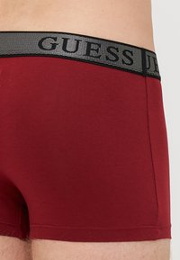 Boxer brevi rossi con un elastico a rete nera che presenta il logo "GUESS." Texture liscia, design aderente e gamba corta.