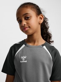 Jeune fille aux cheveux bouclés attachés en arrière, portant un maillot de sport gris et noir avec des accents blancs et un logo Hummel sur la poitrine.