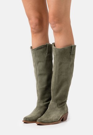 Bottes en daim vert olive à hauteur de genou avec un bout pointu et un talon en bois empilé ; elles présentent une texture douce et un détail replié en haut.