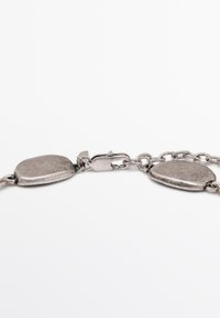 Metallarmbånd med ovale, teksturerede led i sølvfarve og en sikker lås. Designet fremhæver glatte og uregelmæssige former.