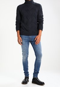 Dunkelblauer Strickpullover mit hohem Kragen und gemustertem Material. Getragen mit hellblauen Skinny-Jeans und schwarzen Stiefeletten.