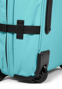 Ein türkisfarbener Koffer mit strukturiertem Stoff, schwarzer Akzent-Hardware, verstärkten Ecken und Doppelfelgen für Mobilität.