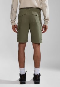 Persona vistiendo pantalones cortos de carga verde oliva, camiseta de manga larga beige, calcetines blancos y zapatos de senderismo negros, de pie sobre un fondo gris claro.