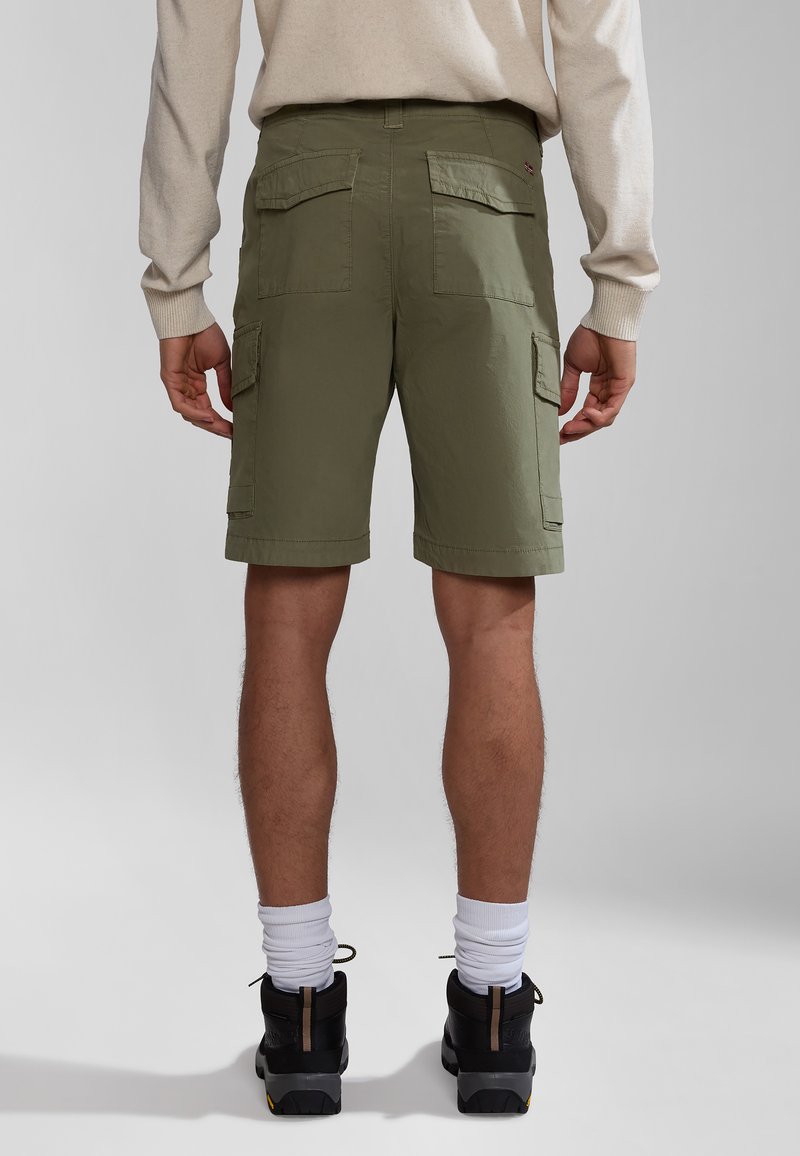 Persona vistiendo pantalones cortos de carga verde oliva, camiseta de manga larga beige, calcetines blancos y zapatos de senderismo negros, de pie sobre un fondo gris claro.