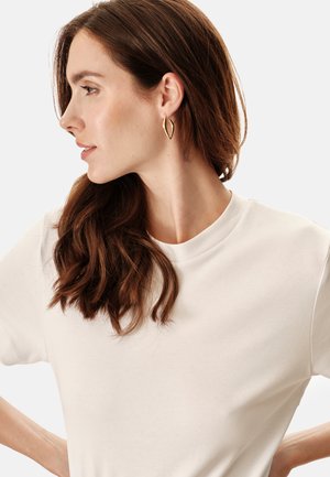 T-shirt crema a girocollo con maniche corte; indossata con orecchini a cerchio a forma di foglia d'oro. Capelli mossi e castani, posizionati sulla spalla.