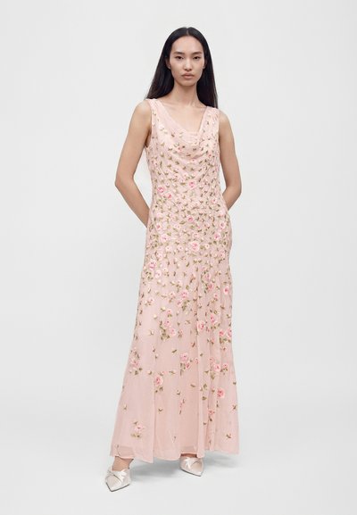 Femme aux longs cheveux foncés portant une robe maxi rose sans manches brodée de fleurs et des chaussures en satin pointues, debout avec les mains dans le dos.