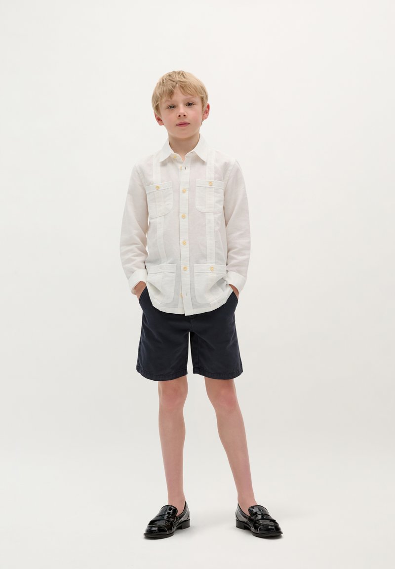 Camicia bianca di lino con bottoni e due tasche frontali, abbinata a pantaloni corti blu scuro e scarpe nere senza lacci. Sfondo semplice.
