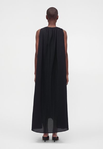 Personne aux cheveux courts portant une robe longue noire sans manches et des talons noirs, debout et de dos sur un fond uni.