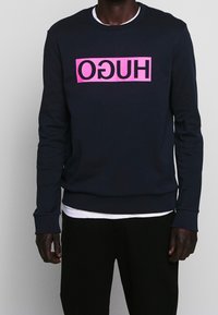 Man som bär en svart sweatshirt med en omvänd rosa rektangulär logotyp med texten "HUGO" på bröstet, över en vit t-shirt och svarta byxor.