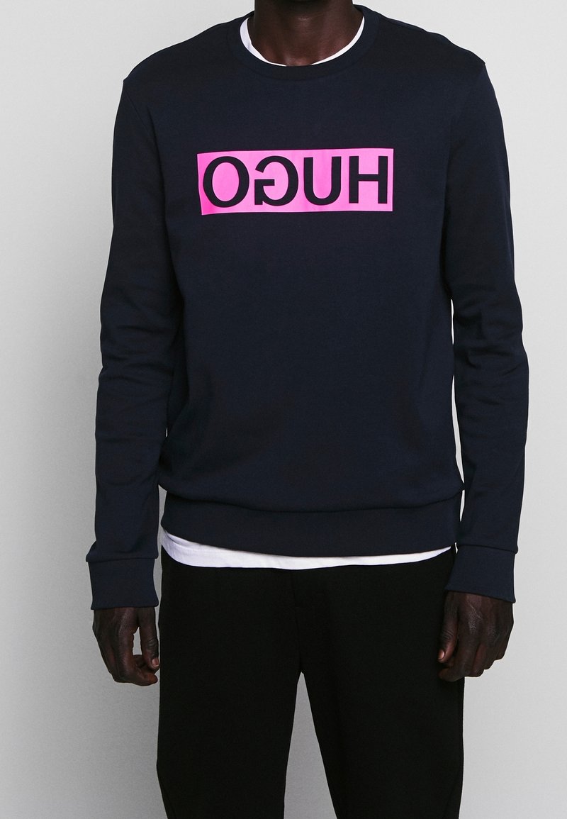 Man som bär en svart sweatshirt med en omvänd rosa rektangulär logotyp med texten "HUGO" på bröstet, över en vit t-shirt och svarta byxor.