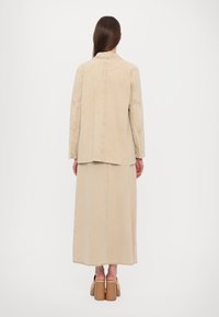 Beige denimjacka med krage och långa ärmar, kombinerad med en matchande golvlång kjol. Båda har en slitna nederkant och sömdetaljer.