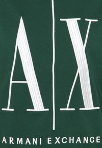Tessuto verde con grandi lettere bianche ricamate "A" e "X", con "ARMANI EXCHANGE" scritto sotto in bianco. Texture liscia.