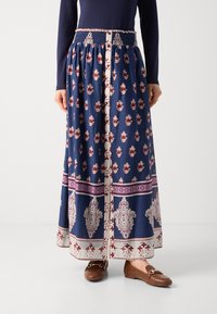 Jupe maxi bleue avec des motifs floraux rouge et blanc, dotée d'une taille smockée et de bords contrastants à l'ourlet. Chaussures plates brunes.