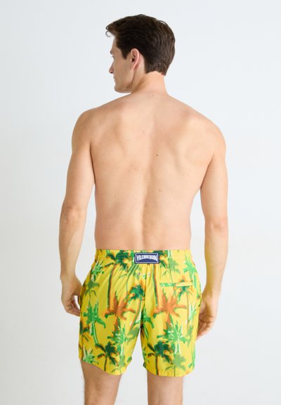 Shorts de bain jaunes avec un motif de palmiers, taille élastique, logo de la marque au dos et poches latérales. Tissu doux avec une texture lisse.
