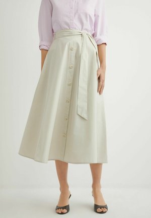 Kobieta ubrana w beżową spódnicę w kształcie litery A zapinaną na guziki, z przewiązanym paskiem w talii, w połączeniu z jasnoróżową koszulą z długim rękawem i czarnymi sandałami z odkrytymi palcami.