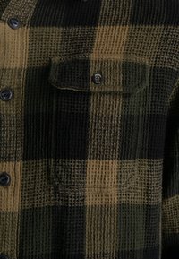 Überprüftes Flanellhemd in Grün, Schwarz und Braun. Verfügt über eine strukturierte Webart, Knopfverschluss und eine Brusttasche mit gebrandeten Beschlägen.