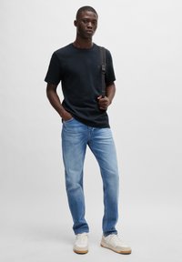 Black cotton t-shirt, light blue jeans, and white sneakers with tan accents. Model standing with hands in pockets and a shoulder strap.