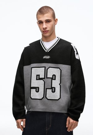 Ung mand med kort hår iført en sort og grå sweater med det hvide strikkede tal 53 på fronten og ærmerne, stående mod en hvid baggrund.