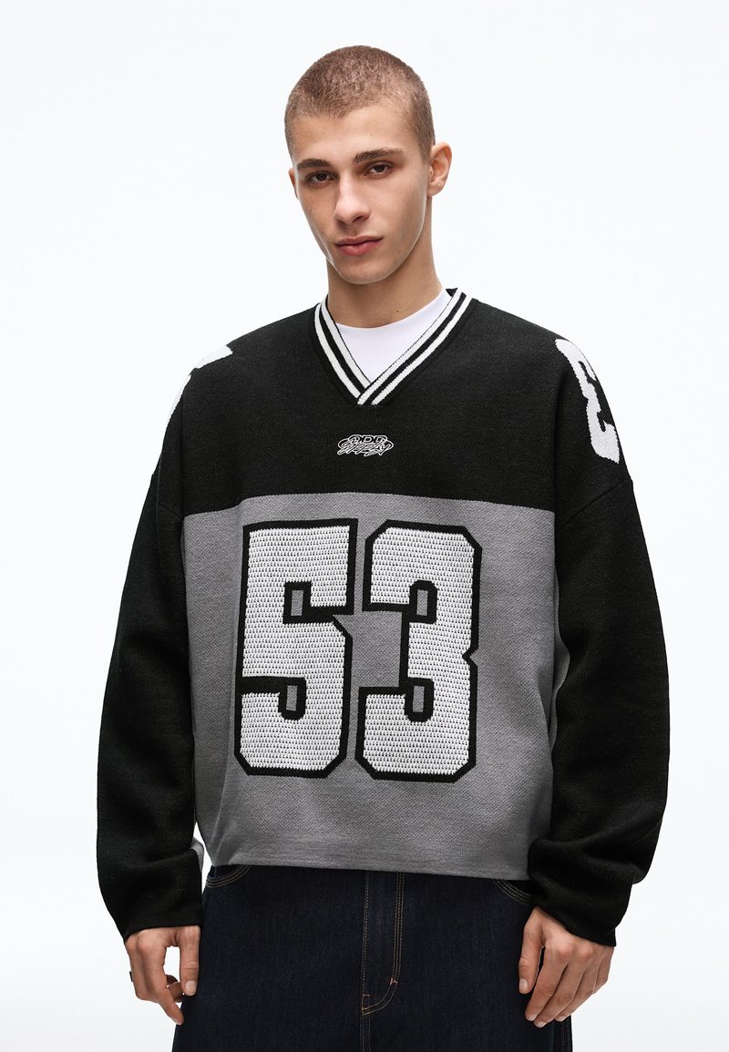 Junger Mann mit kurzen Haaren, der einen schwarzen und grauen Pullover mit der weißen, gestrickten Zahl 53 auf der Vorderseite und den Ärmeln trägt, vor weißem Hintergrund stehend.