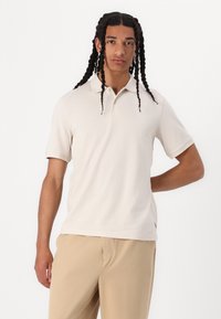 Polo shirt beige a maniche corte con colletto, realizzato in tessuto morbido. Presenta una chiusura con tre bottoni e sottili spacchi laterali all'orlo.