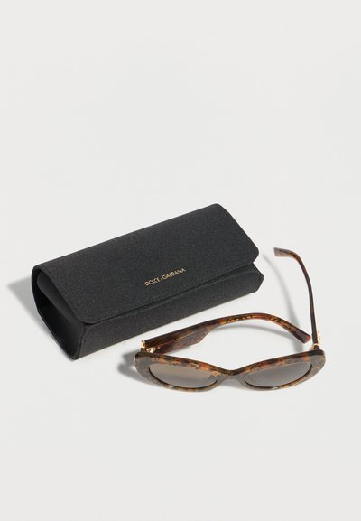 Γυαλιά ηλίου με σκελετό τύπου γάτας σε μοτίβο χελώνας και φακούς με διαβάθμιση χρώματος, δίπλα σε μαύρη υφασμάτινη θήκη με λογότυπο Dolce & Gabbana, πάνω σε λευκή επιφάνεια.