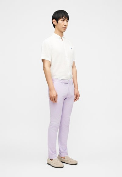 Άνδρας που στέκεται φορώντας λευκό πουκάμισο με κοντά μανίκια και κουμπιά, ανοιχτό μωβ παντελόνι στενής γραμμής και μπεζ μοκασίνια μπροστά σε απλό φόντο.