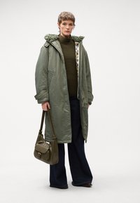 Cappotto lungo verde oliva con cappuccio foderato in pelliccia, abbinato a un maglione scuro e jeans a gamba larga. In mano una borsa verde. Sfondo neutro.