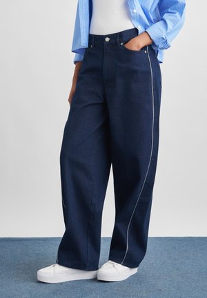 Persoană care poartă blugi bleumarin cu croială largă și tiv alb pe părți, adidași albi, un top alb și o cămașă albastră cu dungi.