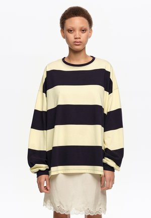 Joven con cabello corto y rizado que lleva una camiseta de manga larga de gran tamaño con amplias rayas horizontales negras y crema y una falda color crema con encaje en el borde.