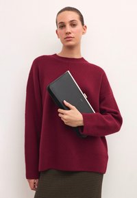 Vynioji burgundiško raudona megztinis su laisvu siluetu ir šonkauliu apvadu, laikoma juoda odine rankine su auksine sagtimi ir lygiu paviršiumi.