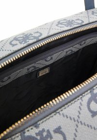 Intérieur d'un sac à main en toile grise à motifs, comportant une poche zippée, une fermeture éclair en métal doré et une étiquette avec le logo "GUESS". Doublure en tissu doux.