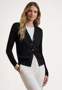 Cardigã preto com design de decote em V, mangas compridas e botões prateados. Usado sobre uma blusa branca, combinado com calças brancas. Textura suave.