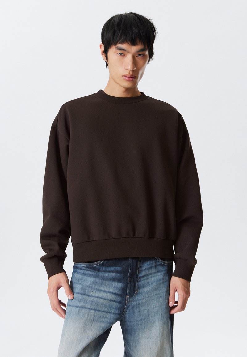 Jeune homme aux cheveux noirs portant un sweat-shirt brun foncé uni et un jean bleu délavé, se tenant contre un fond blanc.