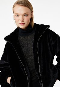 Chaqueta de felpa negra con cremallera y capucha, que presenta una textura suave y una cremallera plateada, superpuesta a una blusa negra transparente.
